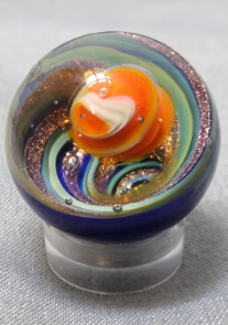 This marble as a smaller orange and white planet floating in the top of a vortex marble. The vortex has sparkly copper, green and blue swirls with a dark blue backing color.